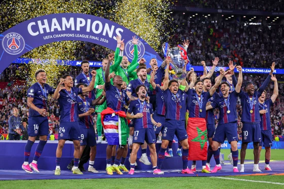 Paris Saint-Germain players celebrate winning the UEFA Champions League Final 2026, lifting the trophy on a podium beneath a "Champions" arch as gold confetti rains down in a packed stadium.